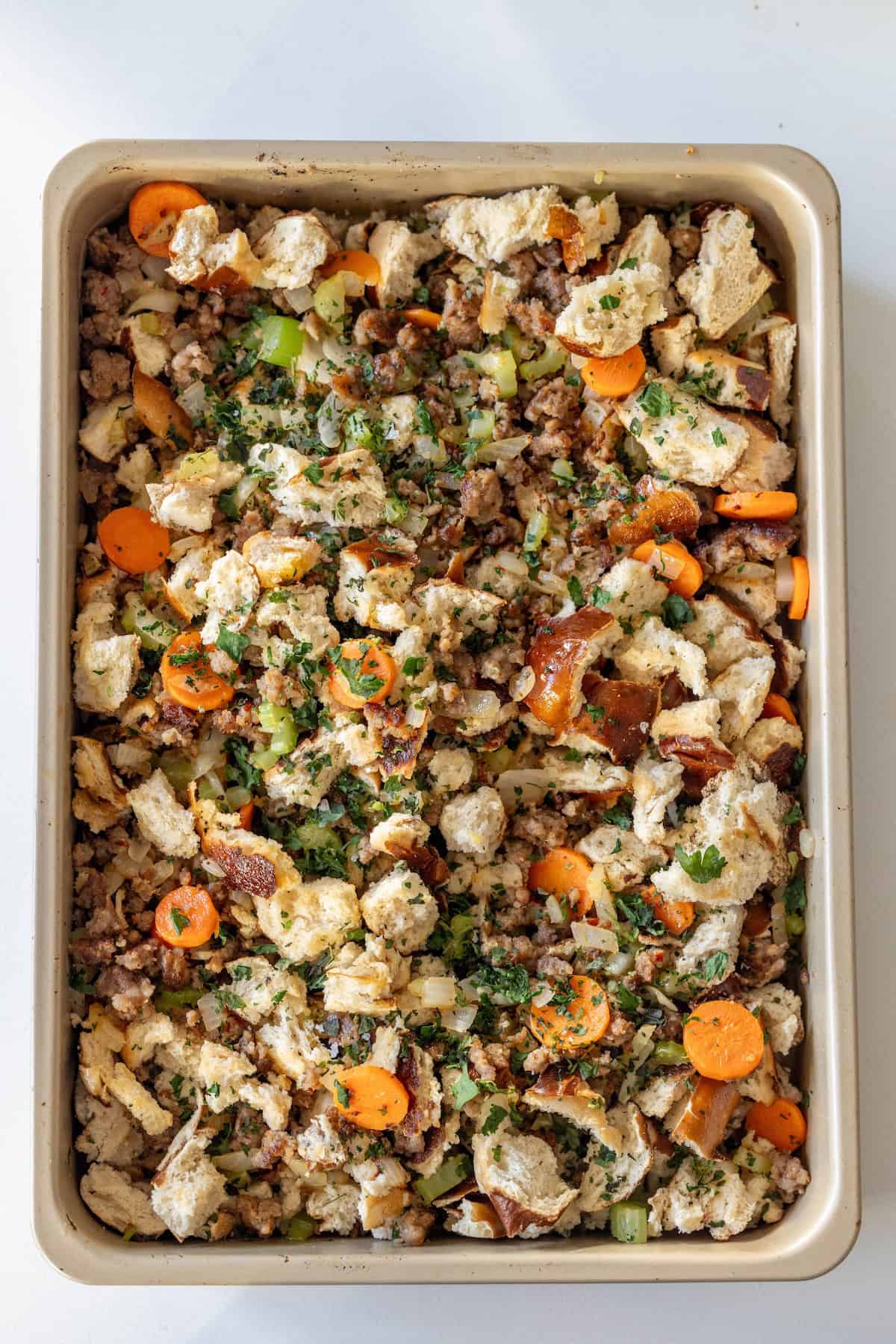 A baking pan filled with unbaked Pretzel Sausage Stuffing mixed with sausage, carrots, celery, and herbs on a white surface.