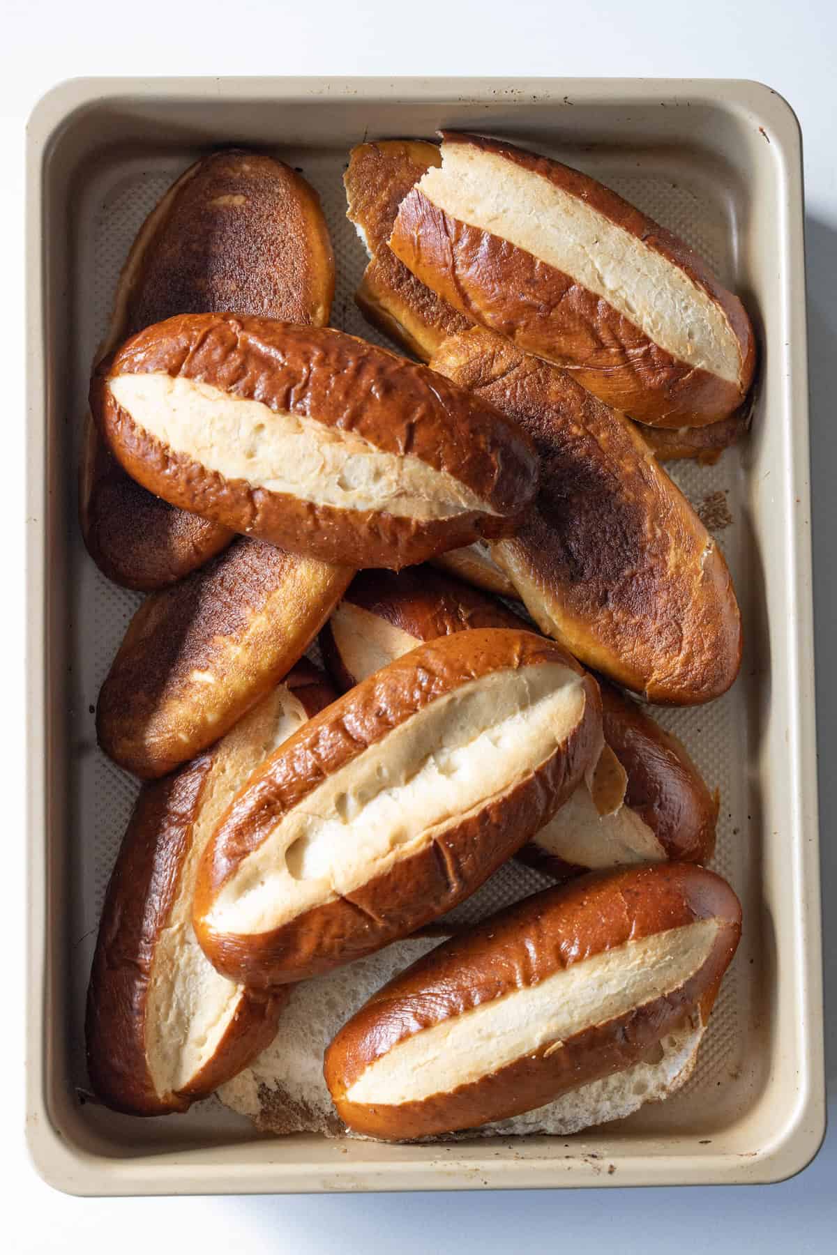 A baking tray filled with several oval-shaped pretzel buns, some lying on their sides and some stacked on top of each other, perfect for making pretzel sausage stuffing.