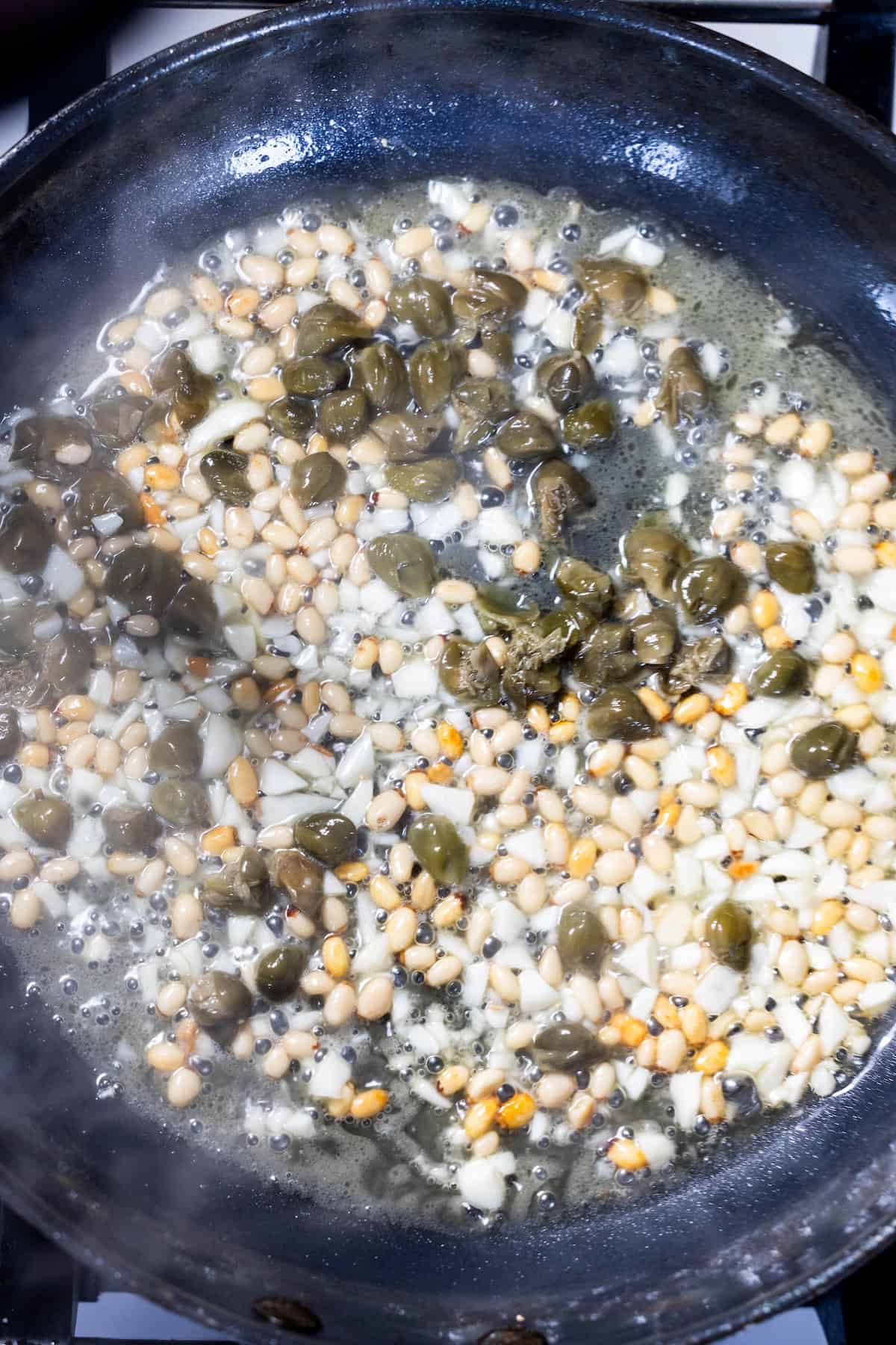 Pine nuts, capers, green olive, and minced garlic are sautéing in a frying pan with oil for a vibrant Puttanesca-inspired flavor.