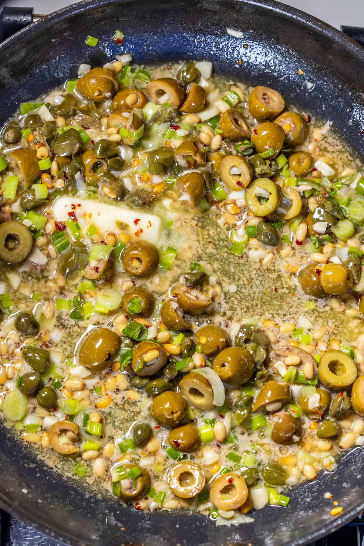 A close-up of a skillet containing green olives, capers, pine nuts, chopped green onions, garlic, red pepper flakes, and butter sizzling in oil—a vibrant mix inspired by classic Puttanesca flavors.