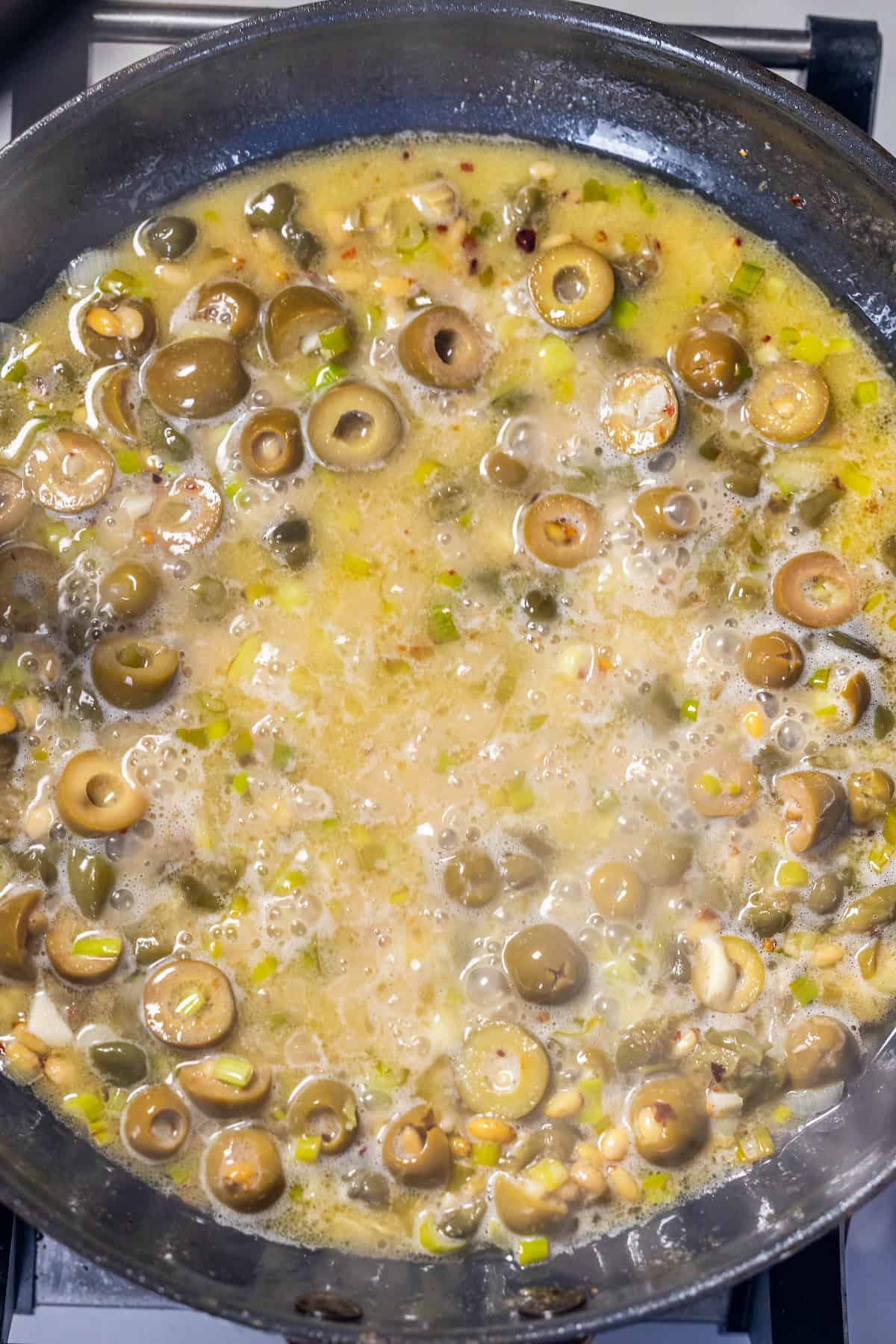 A close-up of a pan on a stovetop with sliced green olives, capers, and other ingredients simmering in a light puttanesca sauce.