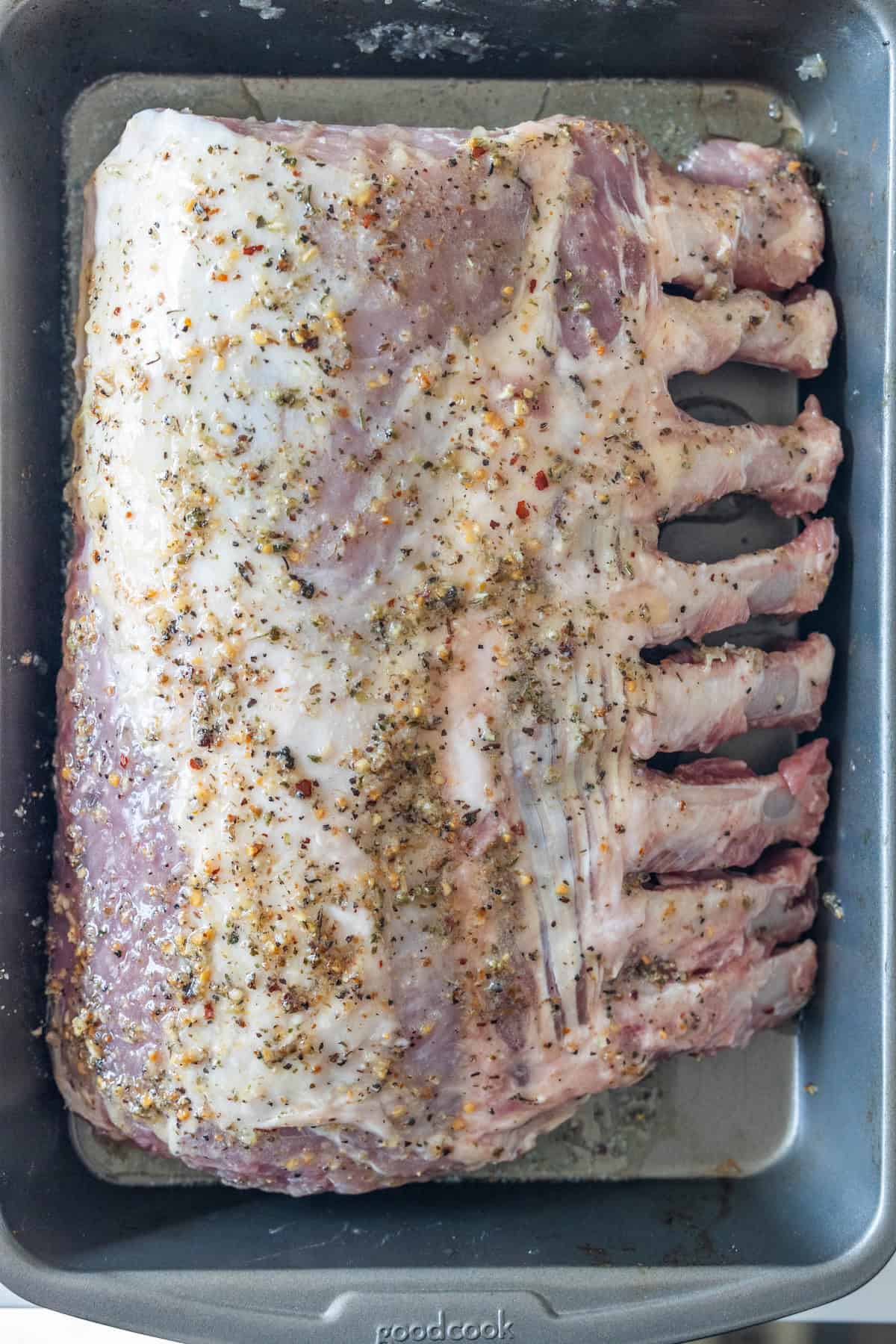 Uncooked rack of lamb coated with a herb and spice marinade, placed in a metal roasting pan—perfect inspiration for your next Roasted Pork Recipe.
