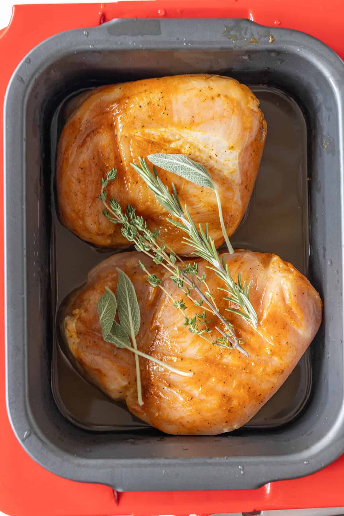 Two seasoned turkey breasts with fresh herbs sit in a roasting pan with liquid, ready for slow cooker preparation.