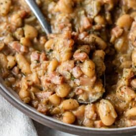 Close-up of brothy ham and beans featuring white beans, diced ham, and herbs in a thick, savory sauce, with a spoon scooping up a portion.