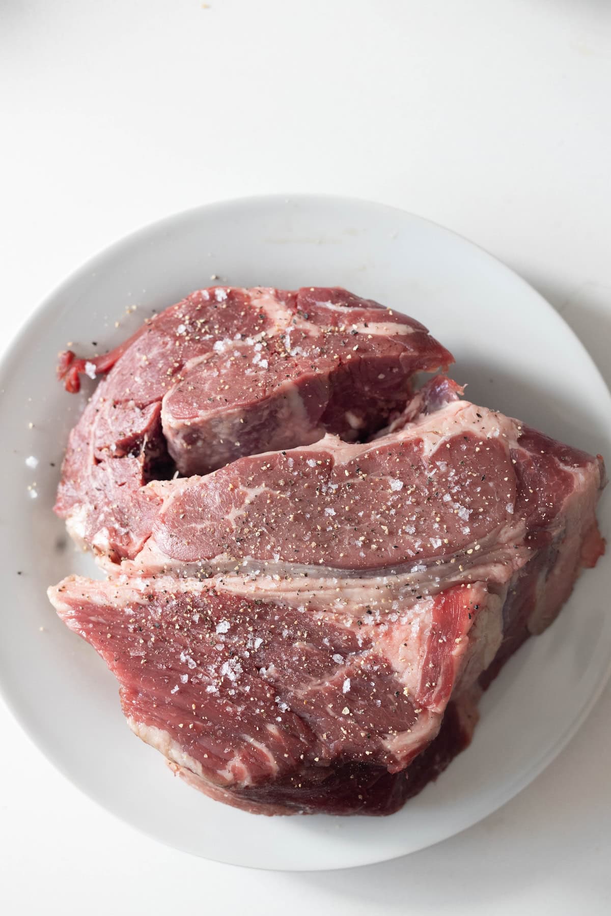 Raw beef chuck roast seasoned with salt and pepper sits on a white plate, ready to become a classic Tomato Soup Pot Roast against a crisp white background.
