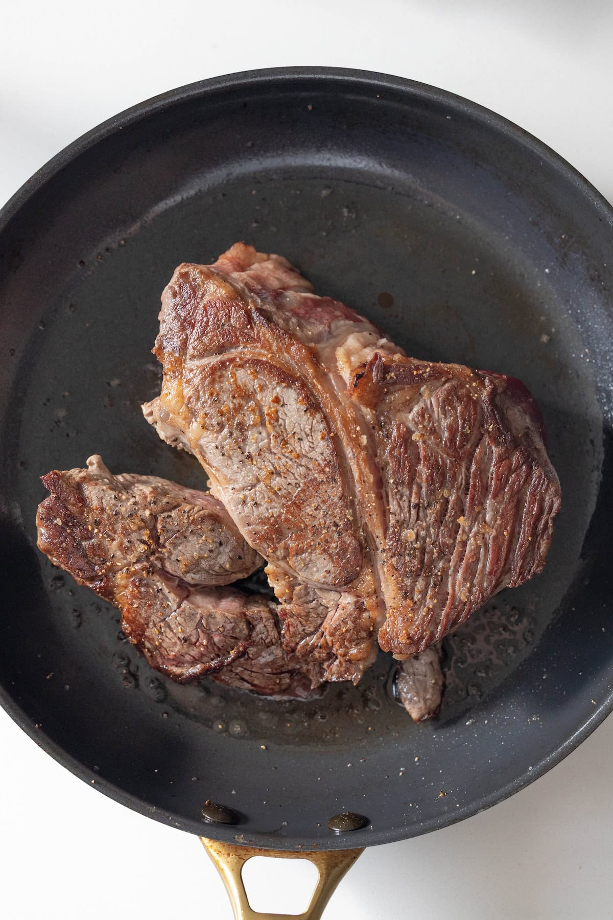 A cooked steak with a browned exterior sits in a black frying pan with a brass handle on a white surface, evoking the hearty aroma of classic Pot Roast.