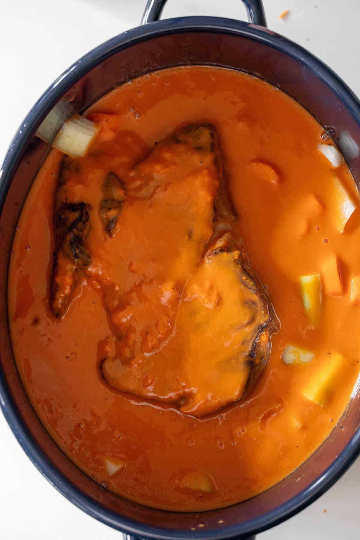 A large pot filled with orange-red sauce, a piece of meat partially submerged, and visible chunks of potatoes or vegetables—this hearty Tomato Soup Pot Roast is comfort food at its finest.