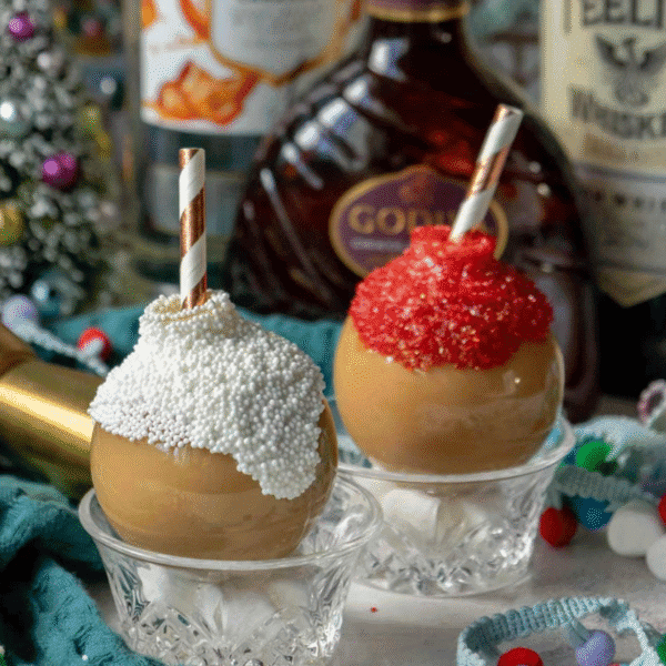 Two caramel-covered apples in glass bowls, topped with white and red sprinkles and striped paper straws, sit beside festive bottles and holiday decor—perfect companions for your favorite holiday drinks.