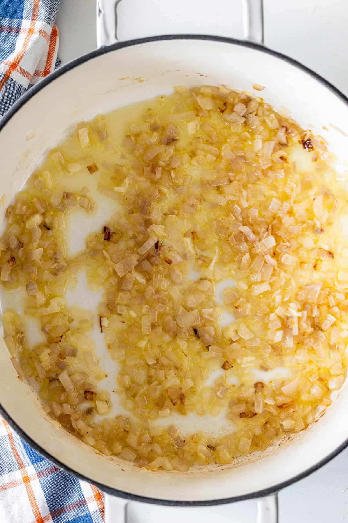 Chopped onions sautéing in a white pot, appearing translucent and lightly browned, with hints of caraway—perfect as a base for brothy ham and beans. A plaid kitchen towel is partially visible in the corner.