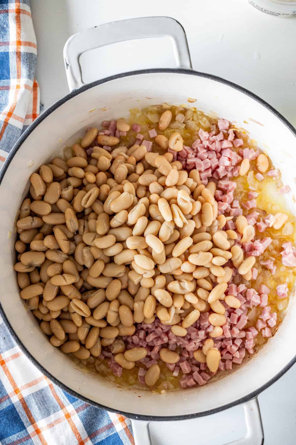 A pot of brothy ham and beans with cooked white beans, chopped ham, and sautéed onions rests beside a blue and orange plaid kitchen towel.