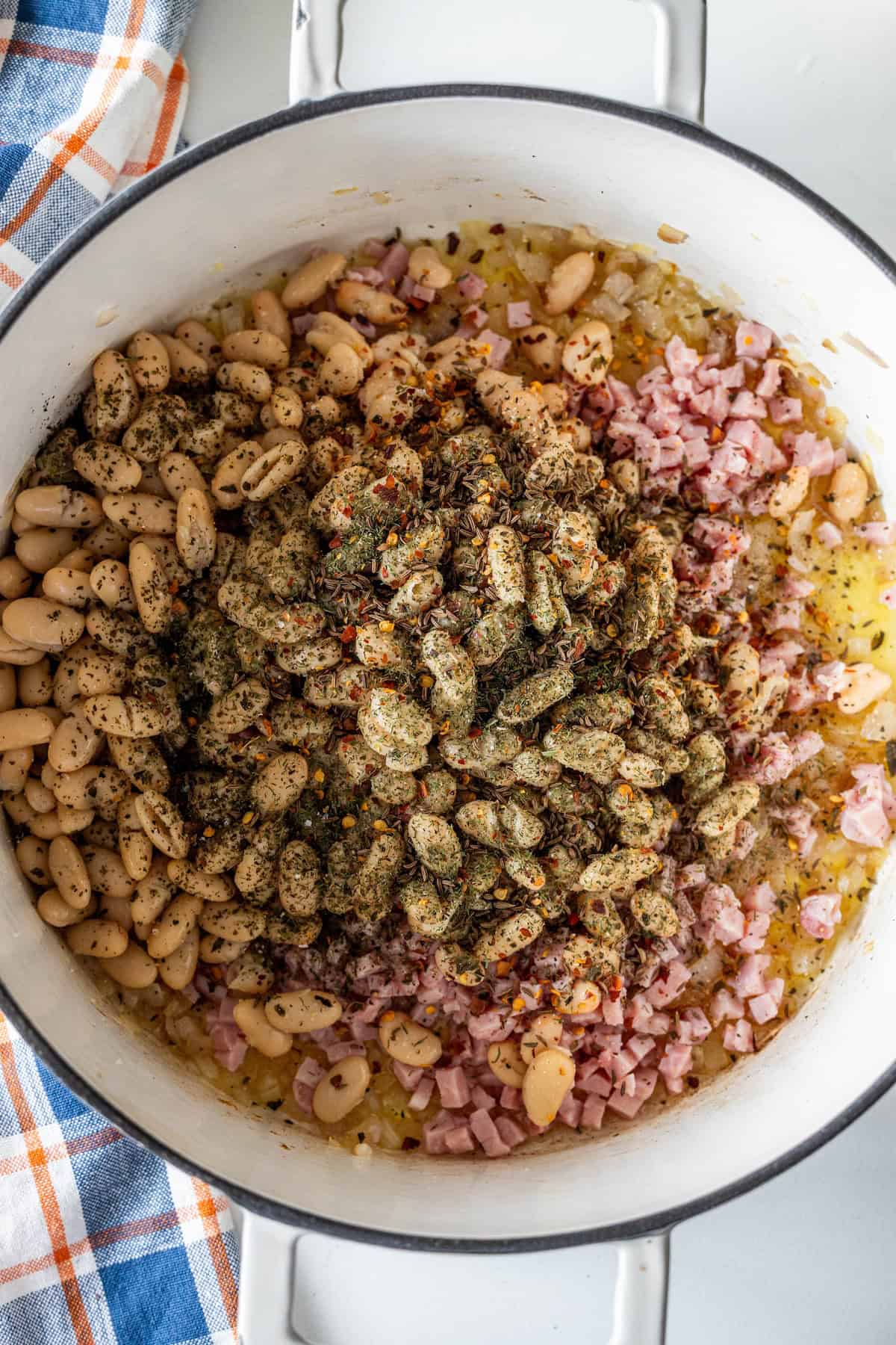 A pot containing white beans, diced ham, chopped onions, and assorted dried herbs and spices, perfect for making brothy ham and beans ready to be mixed and cooked.