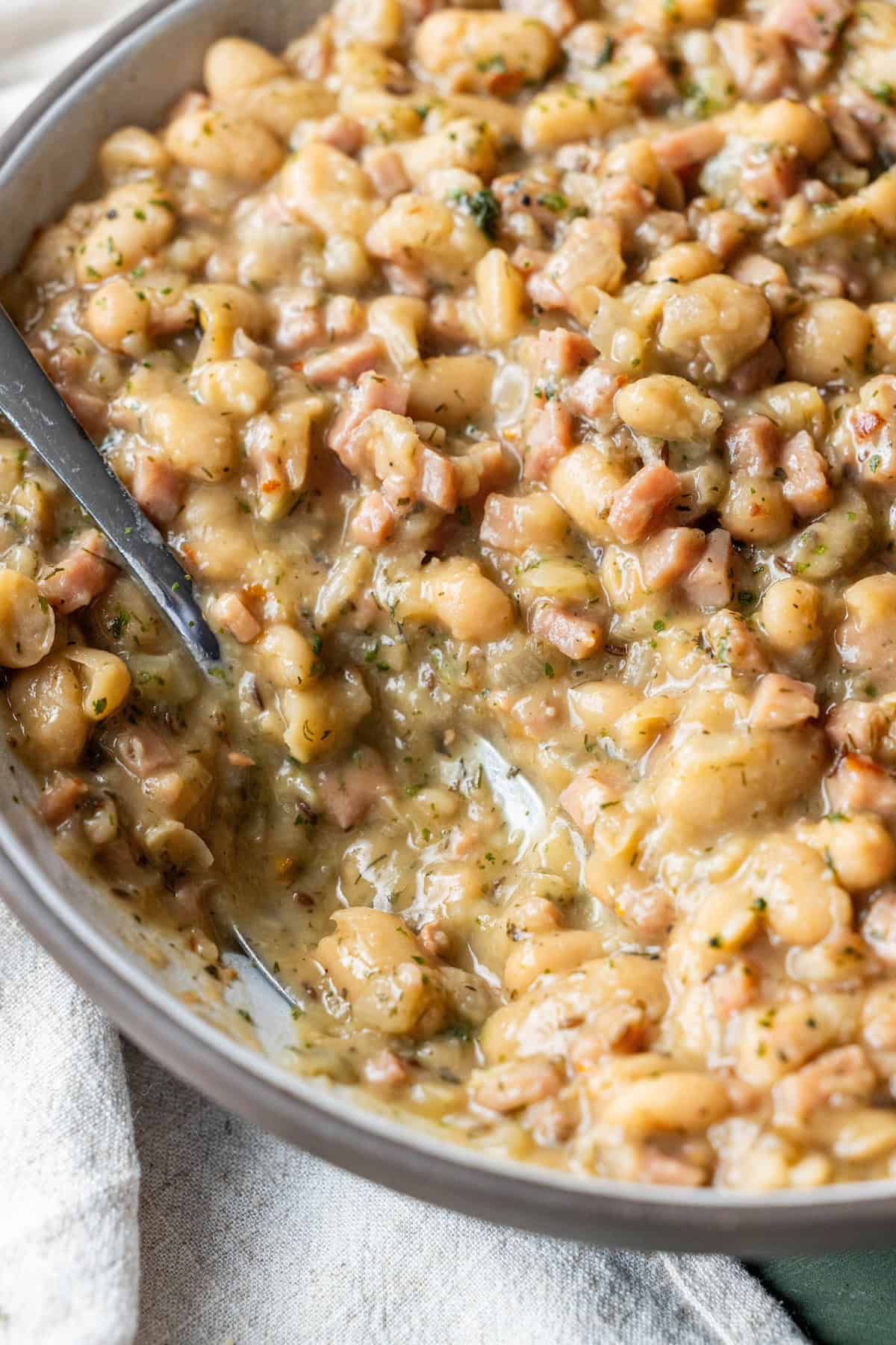 Close-up of a pan filled with brothy ham and beans, creamy white beans mixed with diced ham, fresh herbs, and hints of caraway, with a metal spoon partially submerged in the savory mixture.
