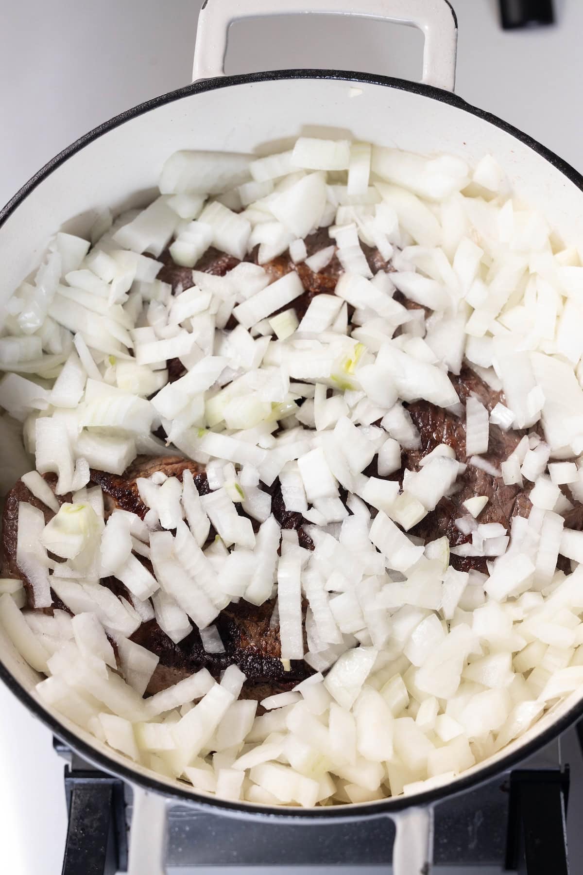 Chopped onions are spread over browned meat pieces in a white pot on a stovetop, perfect for starting a flavorful Onion Soup Pot Roast.