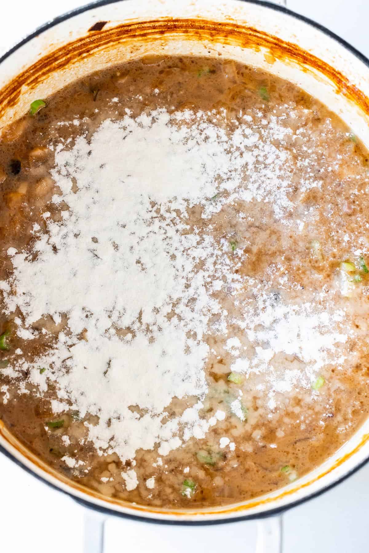A pot of rich onion soup with chopped vegetables, topped with a layer of white flour sprinkled over the surface, reminiscent of a classic Onion Soup Pot Roast.