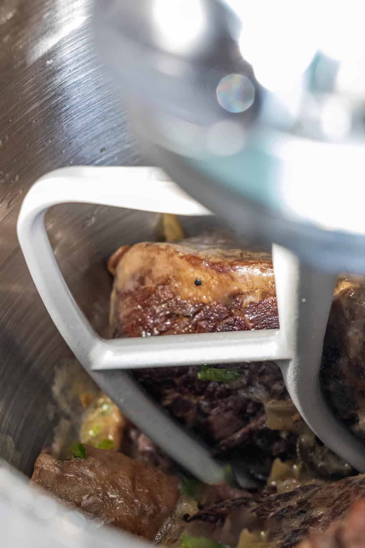 Close-up of beef pieces being mixed in a stand mixer with a metal paddle attachment, perfect for starting a savory Onion Soup Pot Roast.
