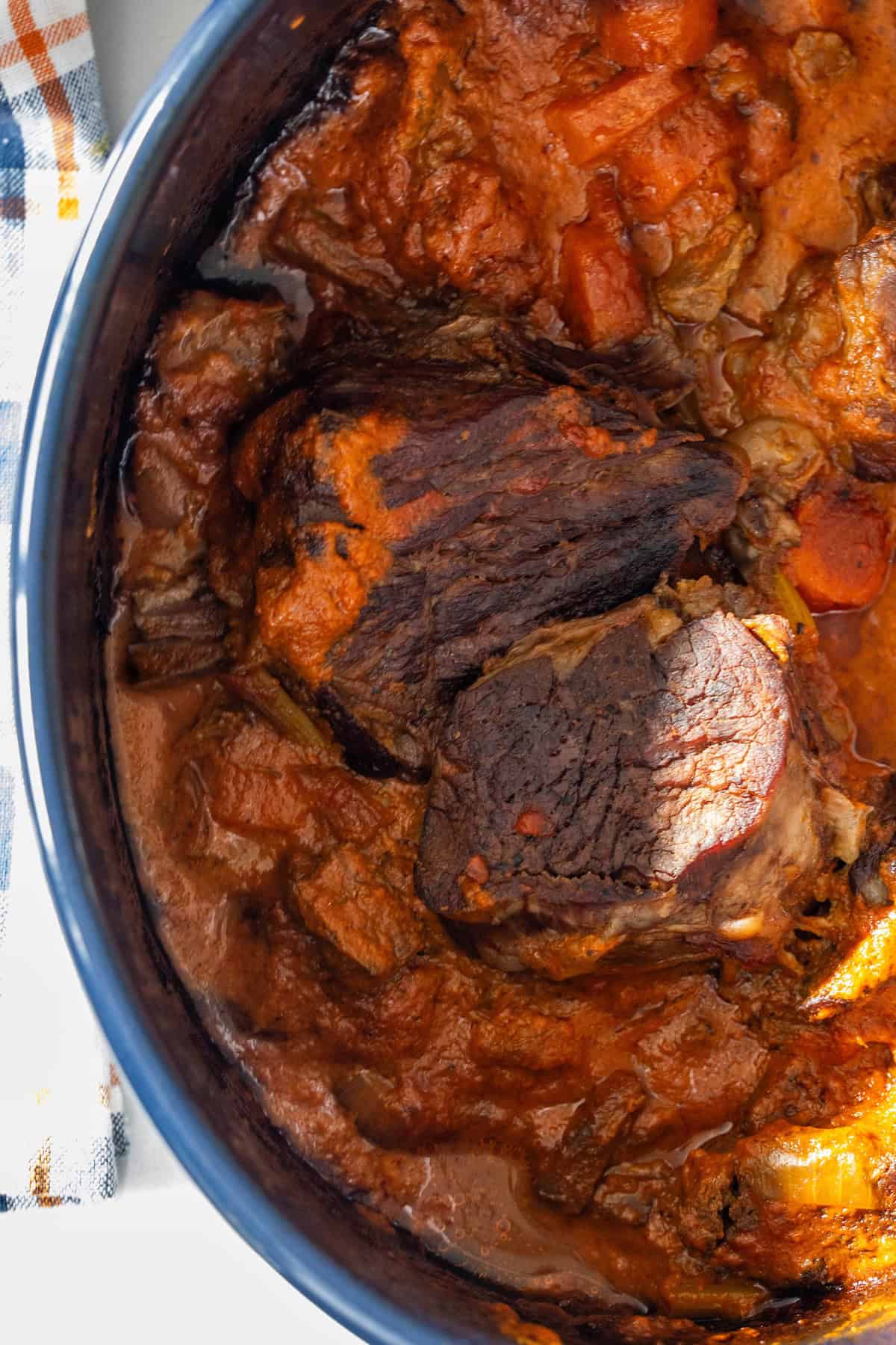 A close-up of Tomato Soup Pot Roast with carrots and savory gravy in a blue Dutch oven, partially bathed in sunlight.