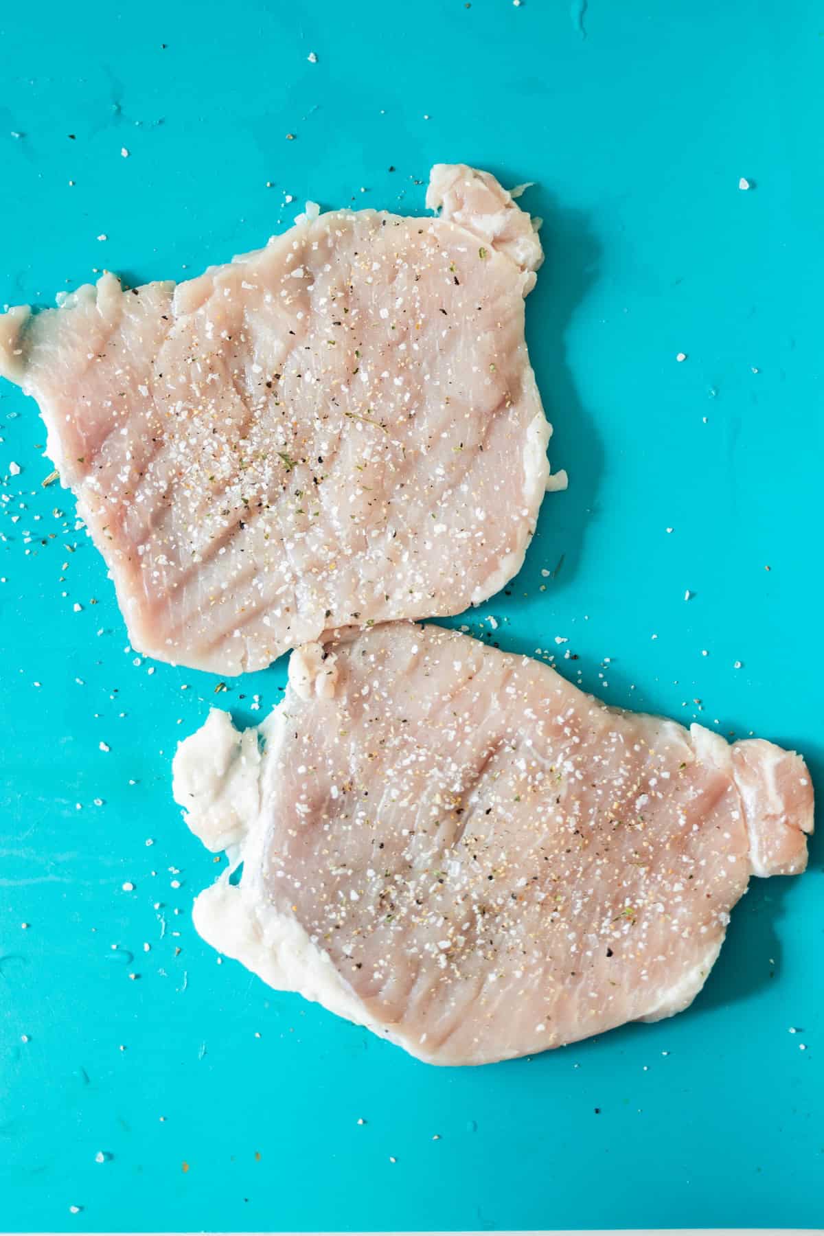 Two raw pork chops, perfect for a Pan Fried Pork Cutlet with Garlic, are seasoned with salt and pepper and placed on a bright turquoise cutting board.