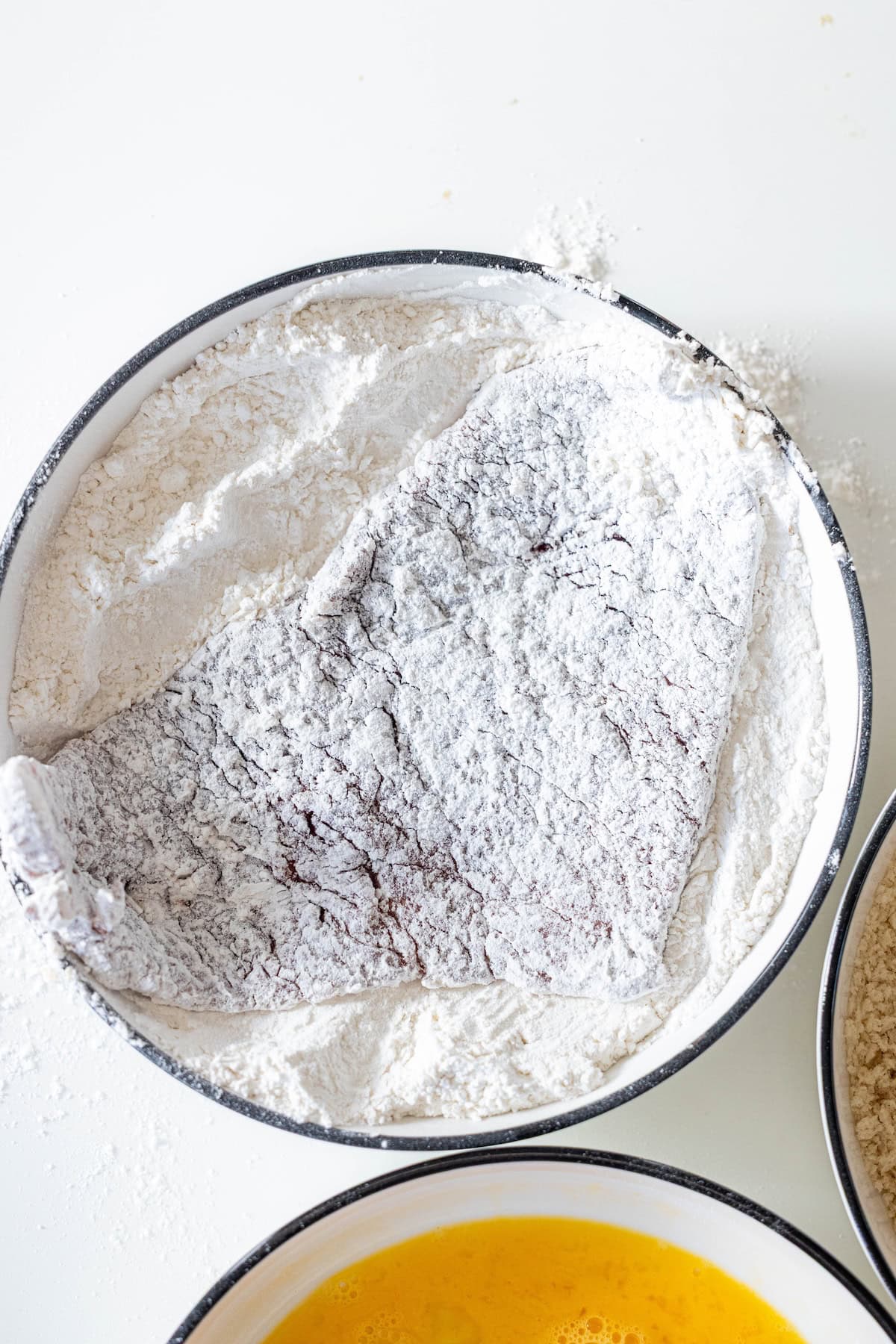 A Steak Milanesa, coated in flour, rests in a white bowl, with the edge of a bowl of beaten eggs visible nearby.