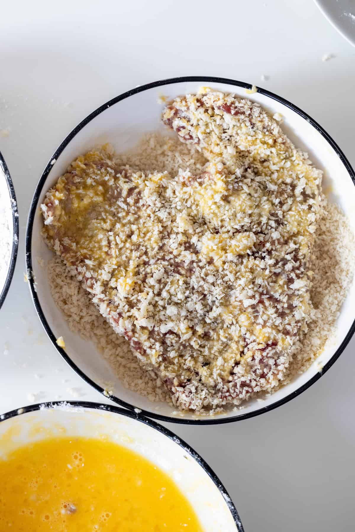 A piece of Steak Milanesa, raw and coated in breadcrumbs, sits in a round white bowl; a bowl with beaten eggs is partially visible beside it.