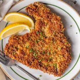 A Steak Milanesa breaded and fried cutlet garnished with chopped chives, served with two lemon wedges on a plate with a fork.