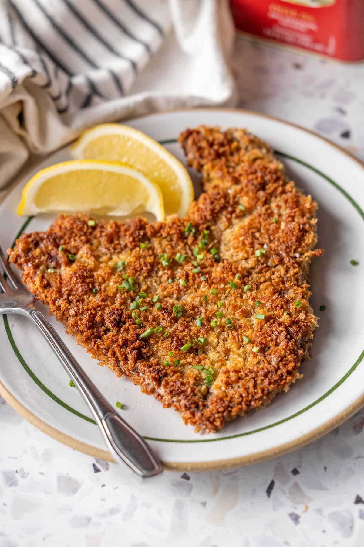 A breaded and fried Milanesa cutlet garnished with chopped herbs is served on a plate with lemon wedges and a fork.
