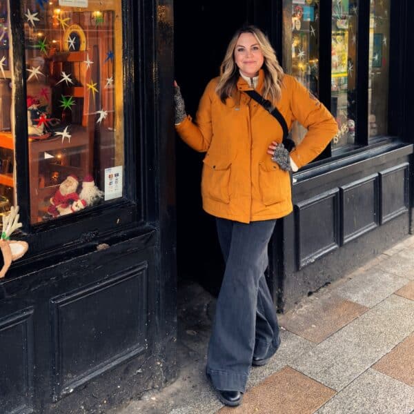 A woman wearing a mustard yellow jacket and dark pants stands in a doorway next to a shop window decorated with colorful stars and festive items.