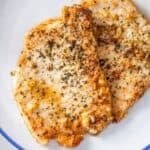 Two seasoned, cooked pork chops on a white plate, reminiscent of pan fried pork cutlet with garlic, sprinkled with herbs and spices.