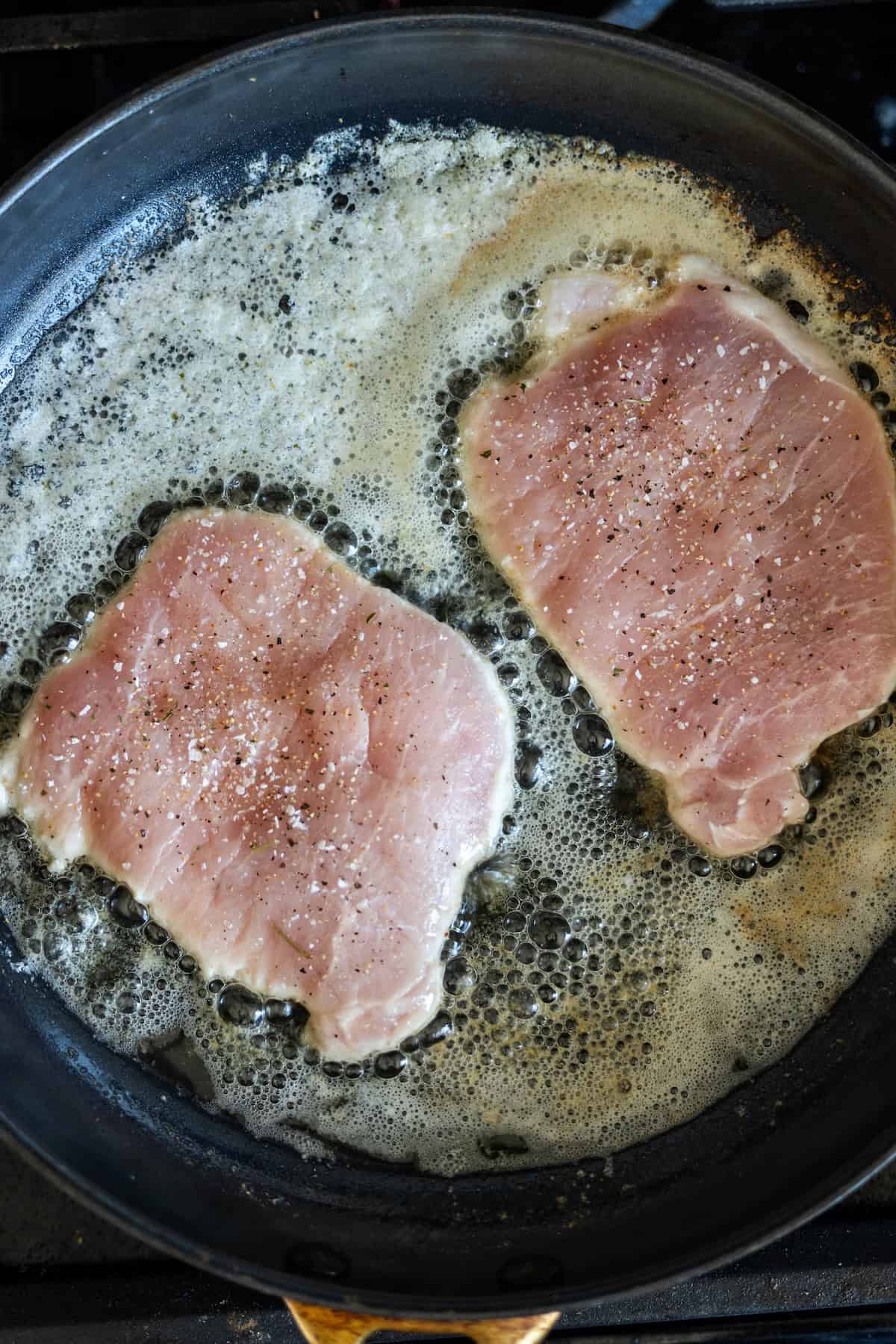 Two raw pork chops, perfect for a pan fried pork cutlet with garlic, are seasoned with salt and pepper as they sizzle in oil and butter in a frying pan.