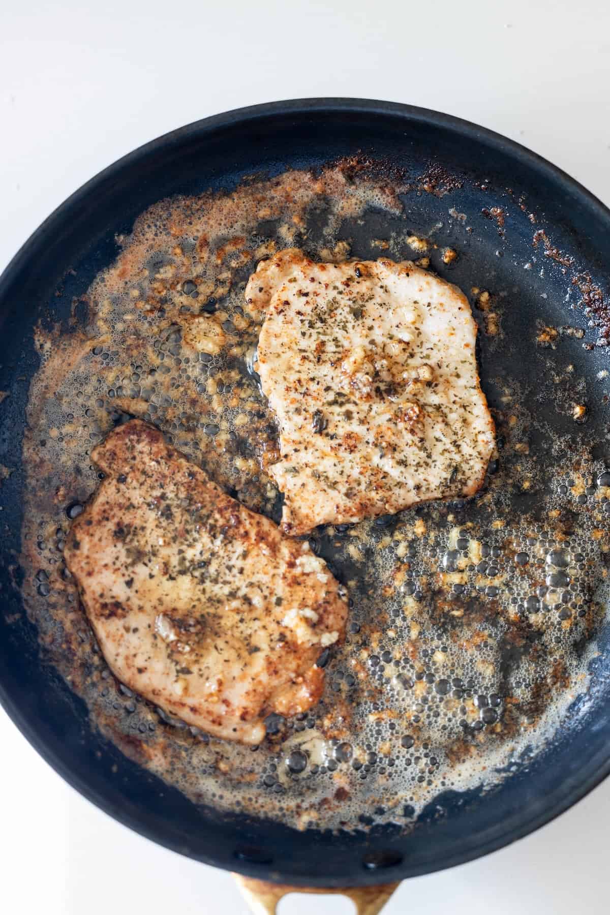 Two seasoned chicken breasts are being cooked in a black skillet, much like a Pan Fried Pork Cutlet with Garlic, with delicious browned bits and savory juices.