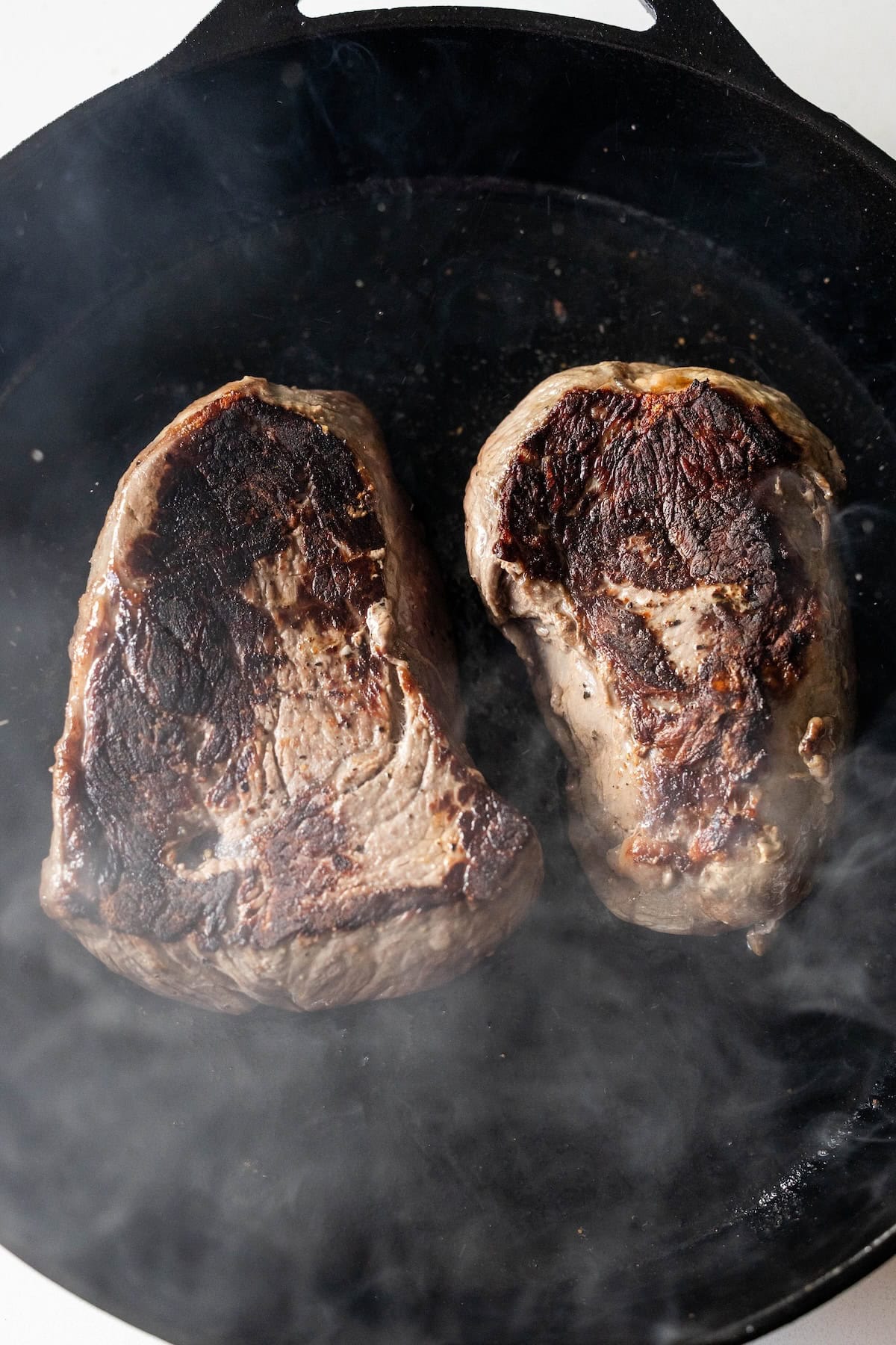 Two pieces of sirloin steak are searing in a black cast iron skillet, with visible browning and some smoke rising around them—capturing the essence of the best sirloin steak cooking experience.