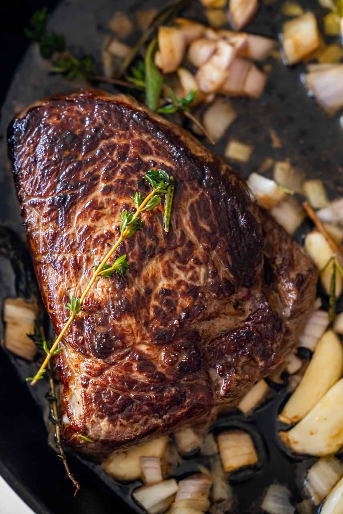 A seared sirloin steak in a pan with chopped onions, garlic cloves, and sprigs of fresh herbs.