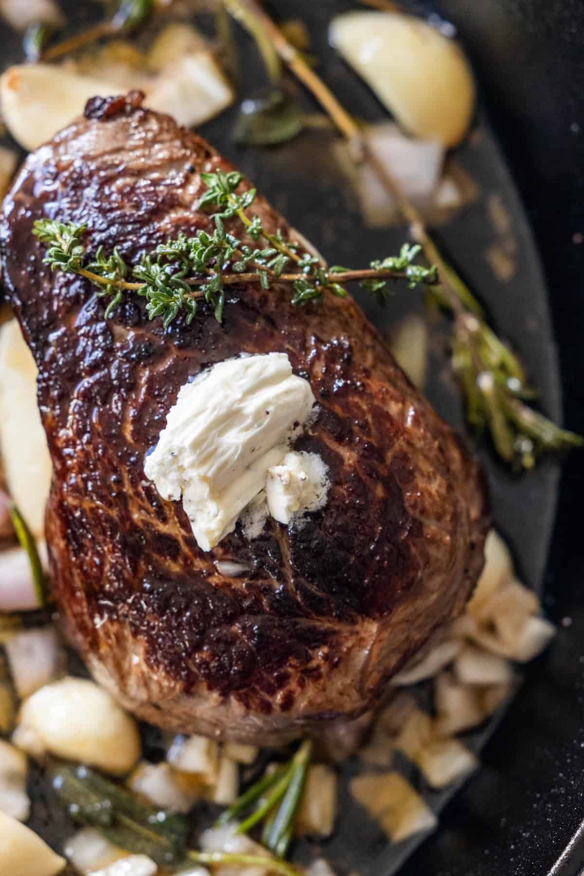 A cooked sirloin steak topped with a dollop of butter and a sprig of thyme, surrounded by garlic herb cloves and fresh herbs in a pan.
