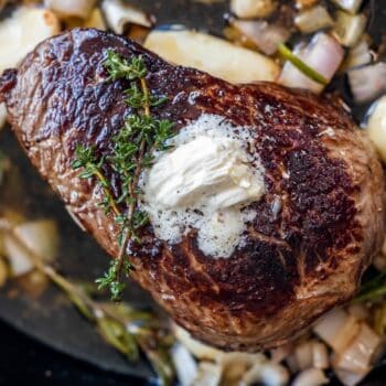 A seared sirloin steak topped with a dollop of butter and a sprig of thyme, surrounded by chopped onions, garlic, and fresh herbs in a pan.