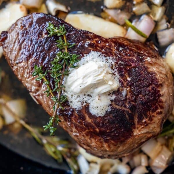 A seared sirloin steak topped with a dollop of butter and a sprig of thyme, surrounded by chopped onions, garlic, and fresh herbs in a pan.