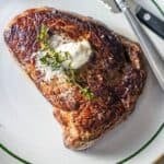 A cooked sirloin steak topped with a dollop of butter and a sprig of thyme on a white plate with a green rim, with a fork and knife beside it.