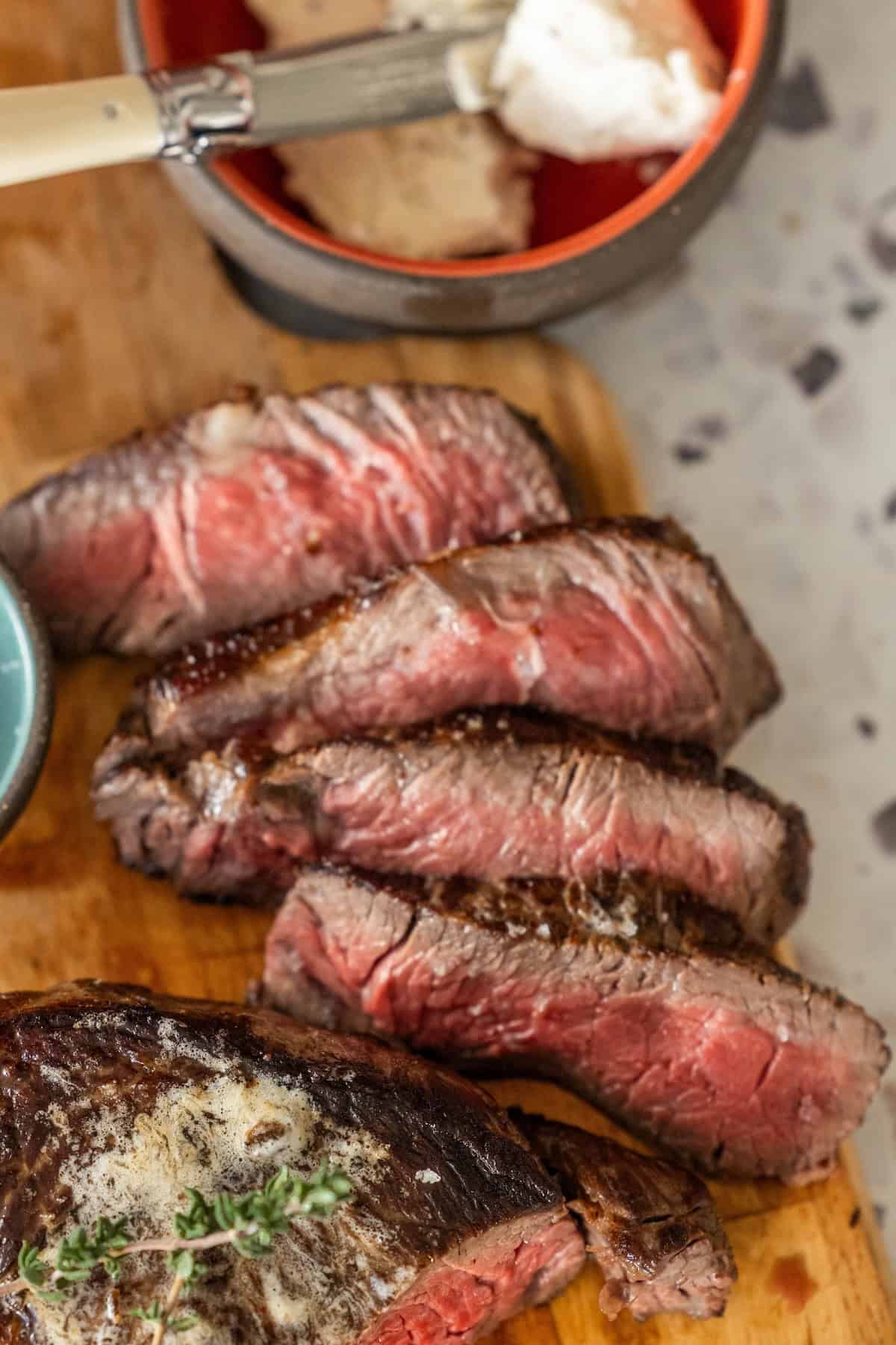 Sliced medium-rare sirloin steak on a wooden board, with a knife and a bowl of creamy garlic herb sauce in the background.