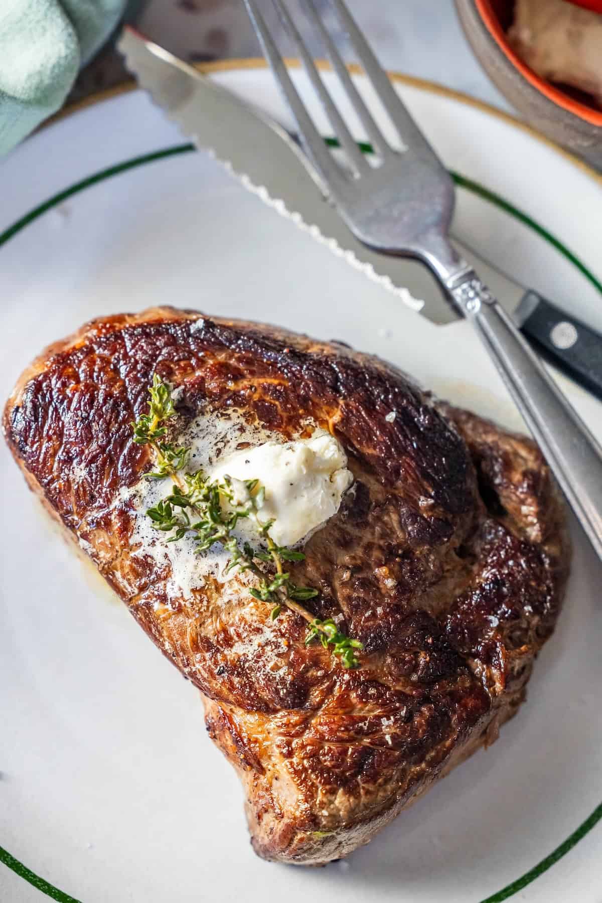A cooked sirloin steak topped with a dollop of butter and a sprig of thyme, served on a white plate with a fork and knife beside it.