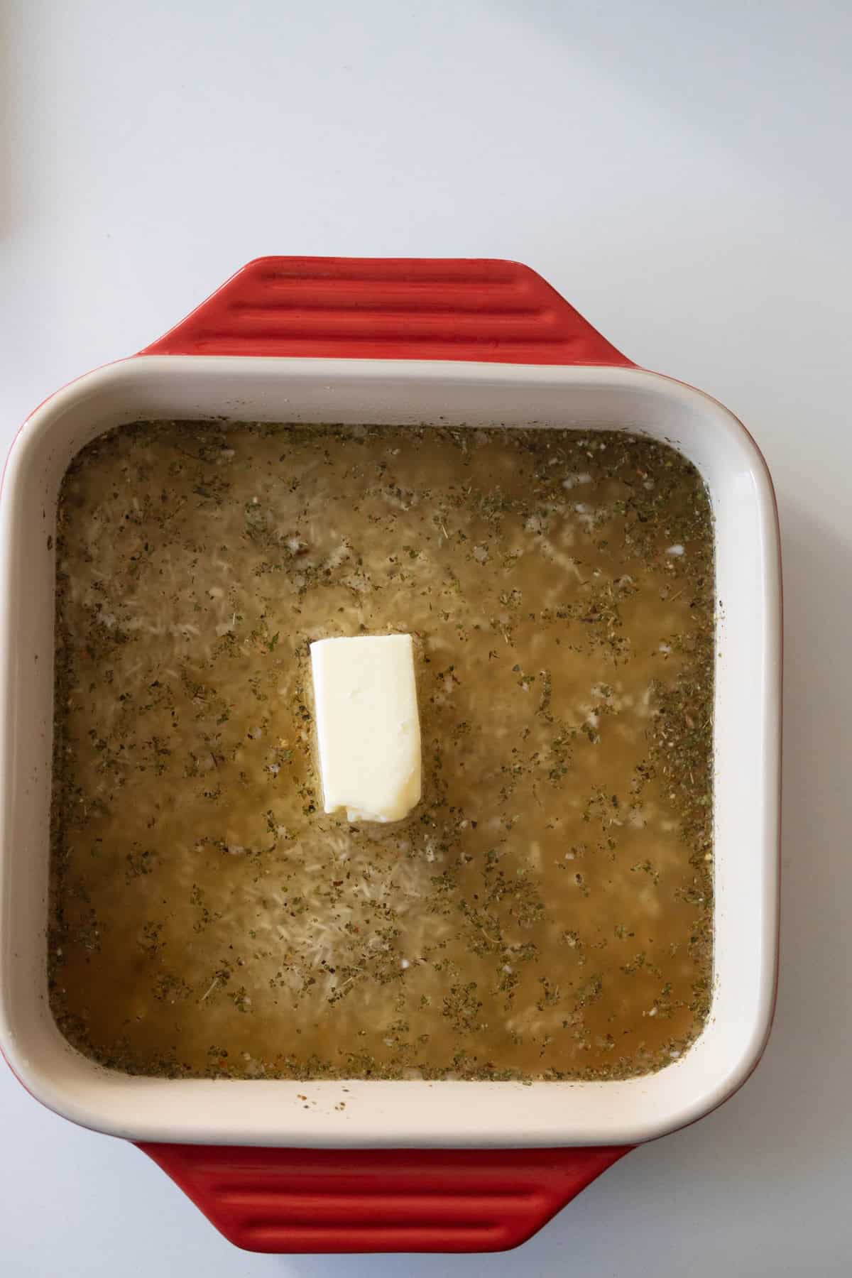 A square red baking dish filled with seasoned broth and a rectangular piece of butter in the center, perfect for making baked lemon rice.