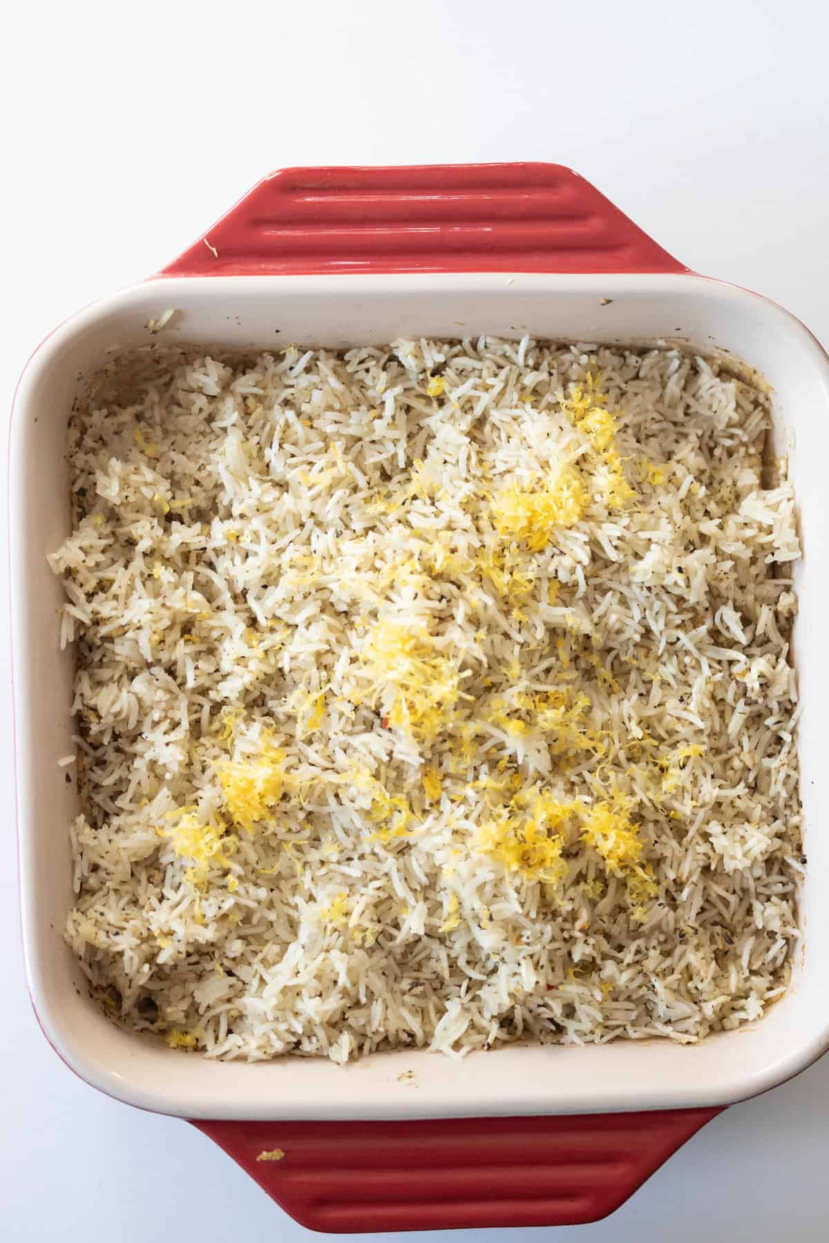 A square red and white baking dish filled with baked lemon rice, topped with fresh lemon zest, sits on a white surface.