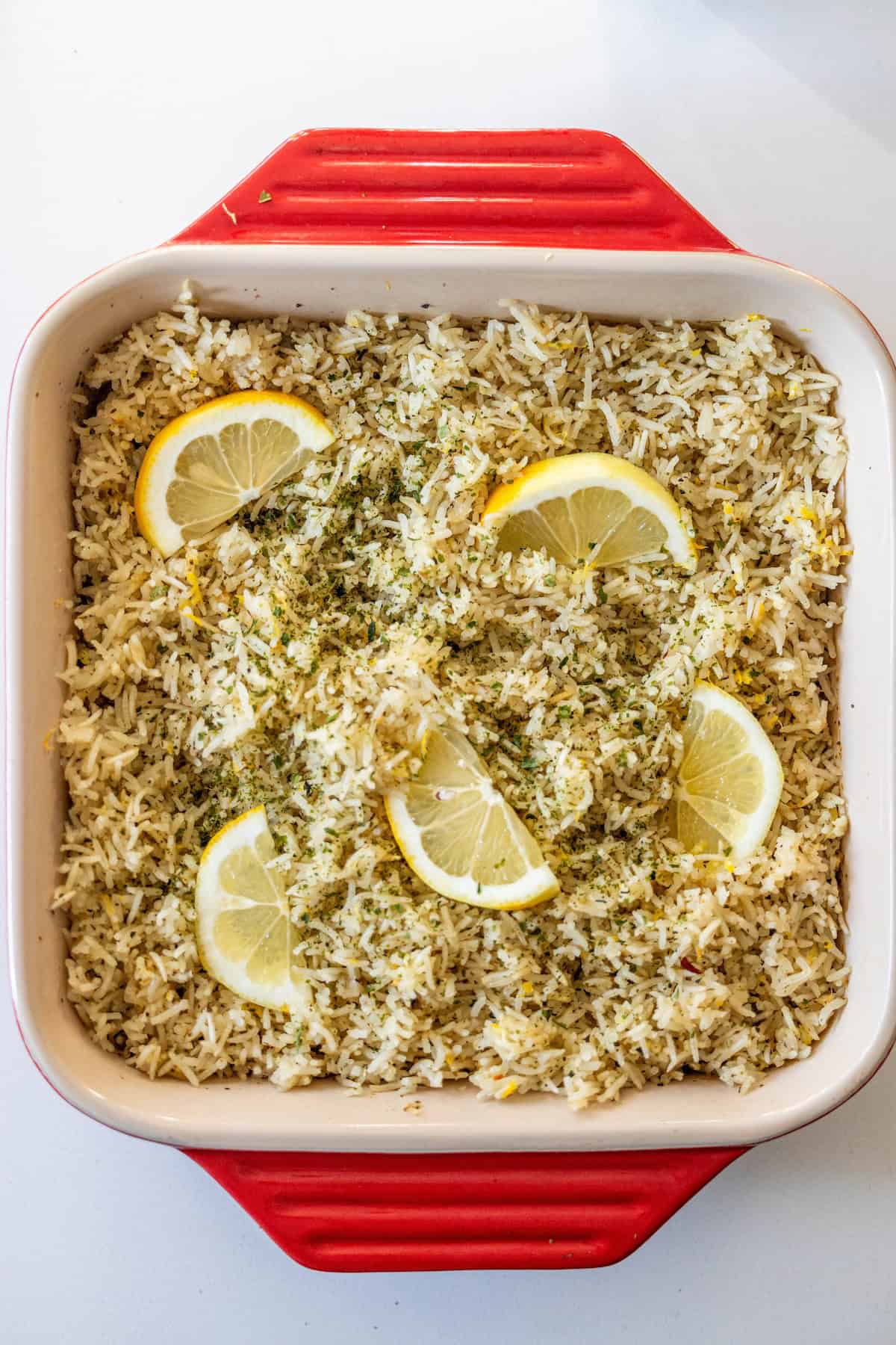 A square red baking dish filled with Baked Lemon Rice, garnished with fresh lemon slices and green herbs.