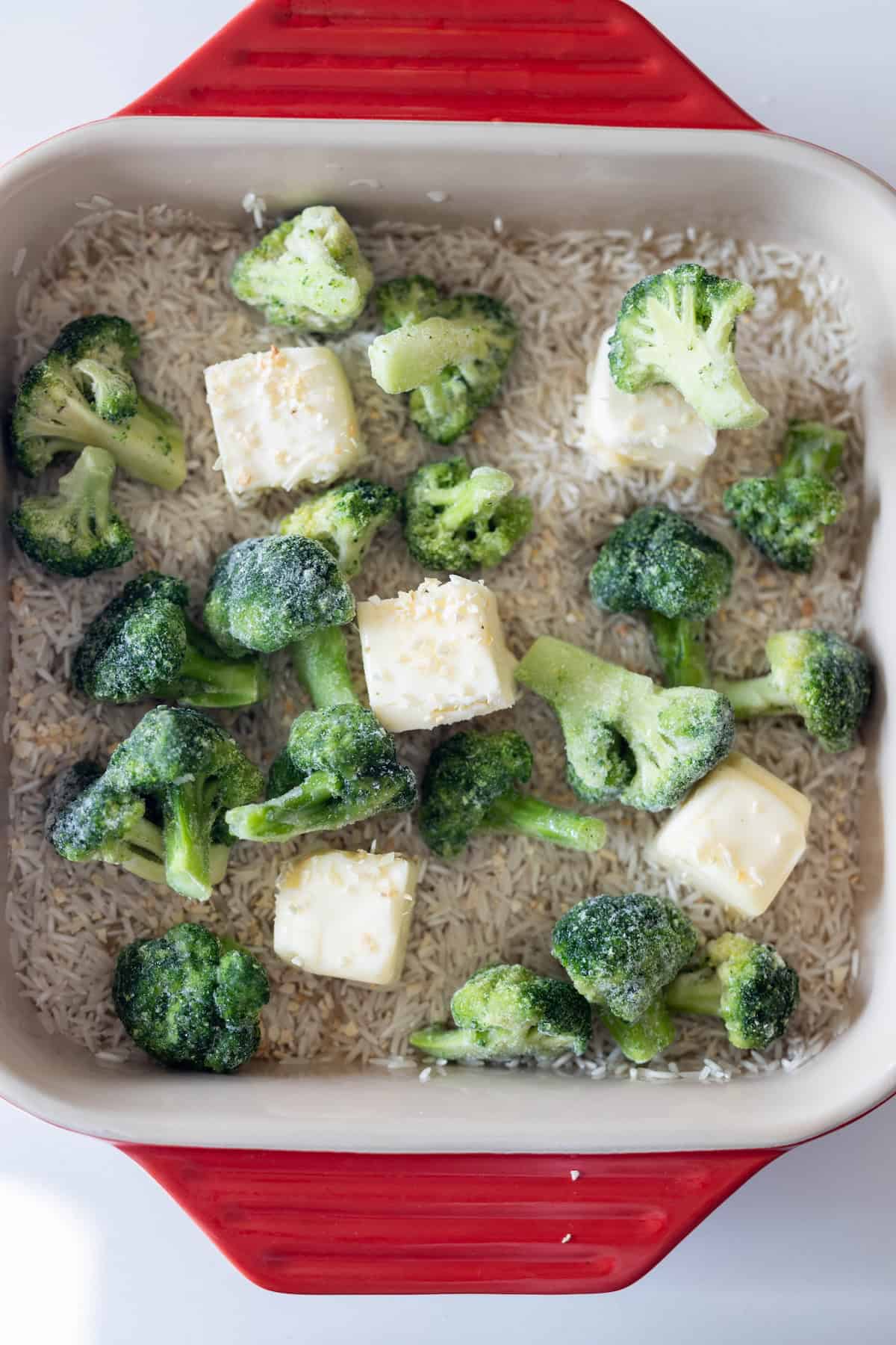 A red baking dish containing uncooked rice, frozen broccoli florets, and cubes of butter, sprinkled with seasoning—perfect for an easy Baked Ranch Rice Recipe.