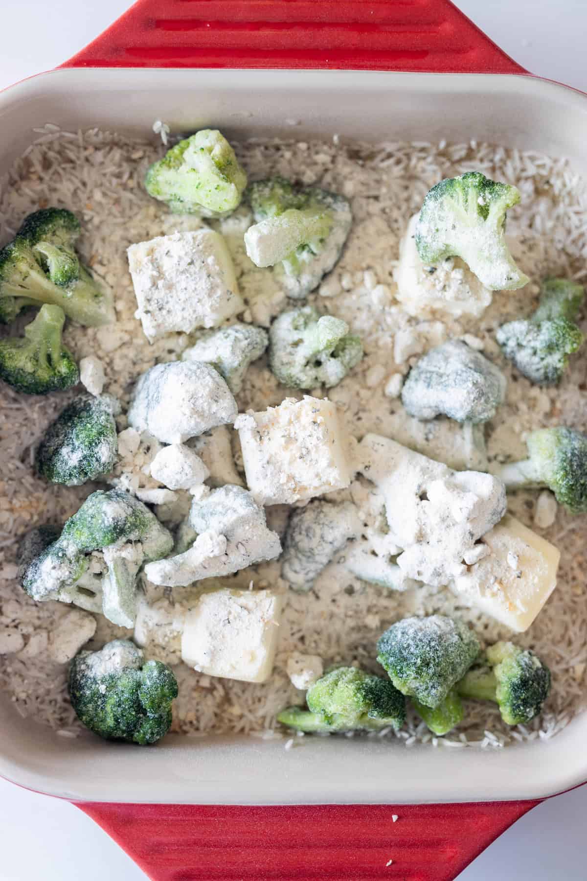 Uncooked rice, frozen broccoli florets, and chunks of butter topped with ranch seasoning in a red and white baking dish—perfect for making Baked Ranch Rice with Broccoli.