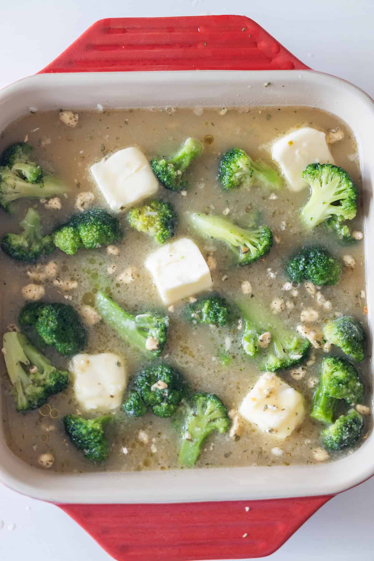 A red-edged baking dish contains broccoli florets and cubes of white cheese in a seasoned liquid, ready to be baked to perfection.
