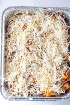 A foil tray filled with cheesy baked rigatoni pasta, topped with shredded cheese and herbs, ready to be baked.