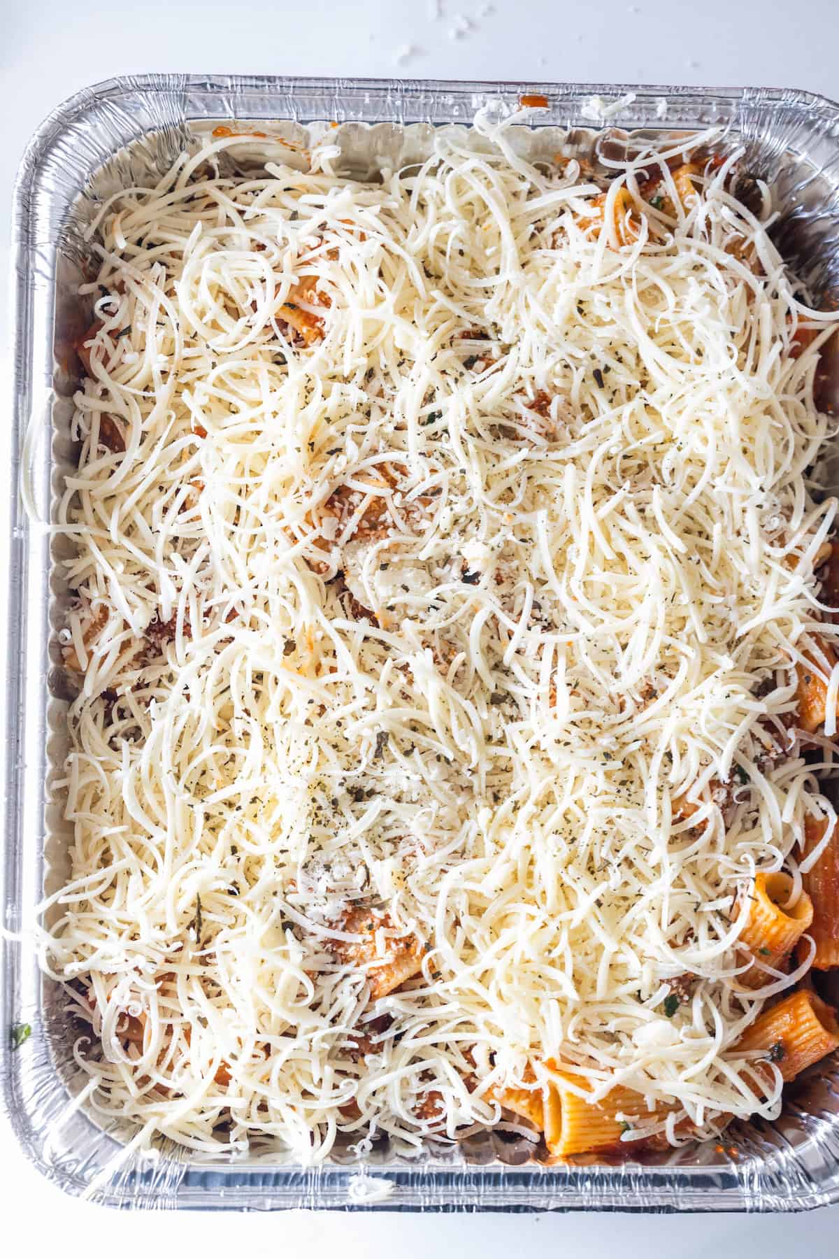 A foil tray filled with cheesy baked rigatoni pasta, topped with shredded cheese and herbs, ready to be baked.