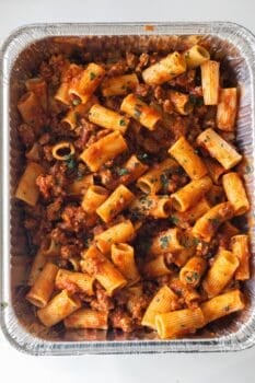 A large aluminum tray filled with cheesy baked rigatoni pasta, topped with ground meat and tomato sauce, and garnished with chopped herbs.