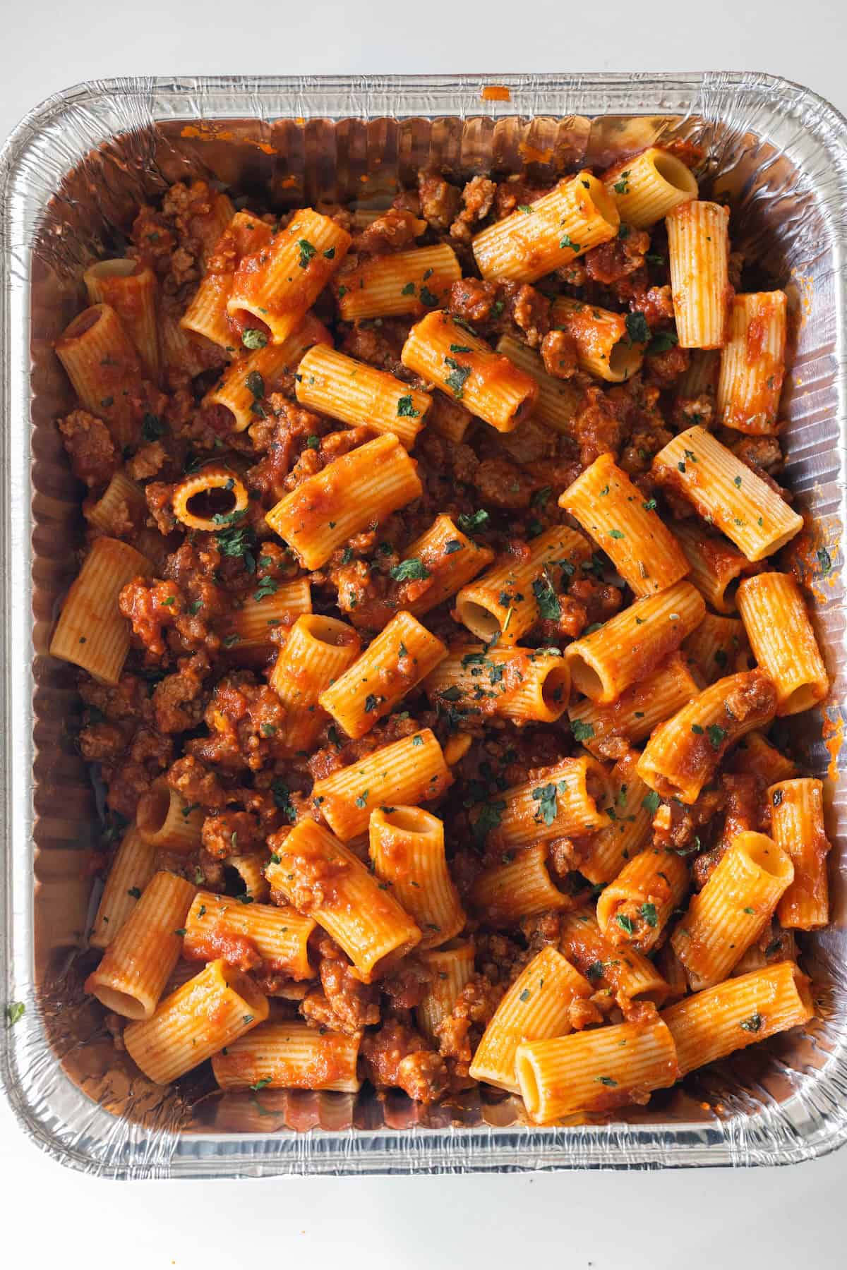 A large aluminum tray filled with cheesy baked rigatoni pasta, topped with ground meat and tomato sauce, and garnished with chopped herbs.