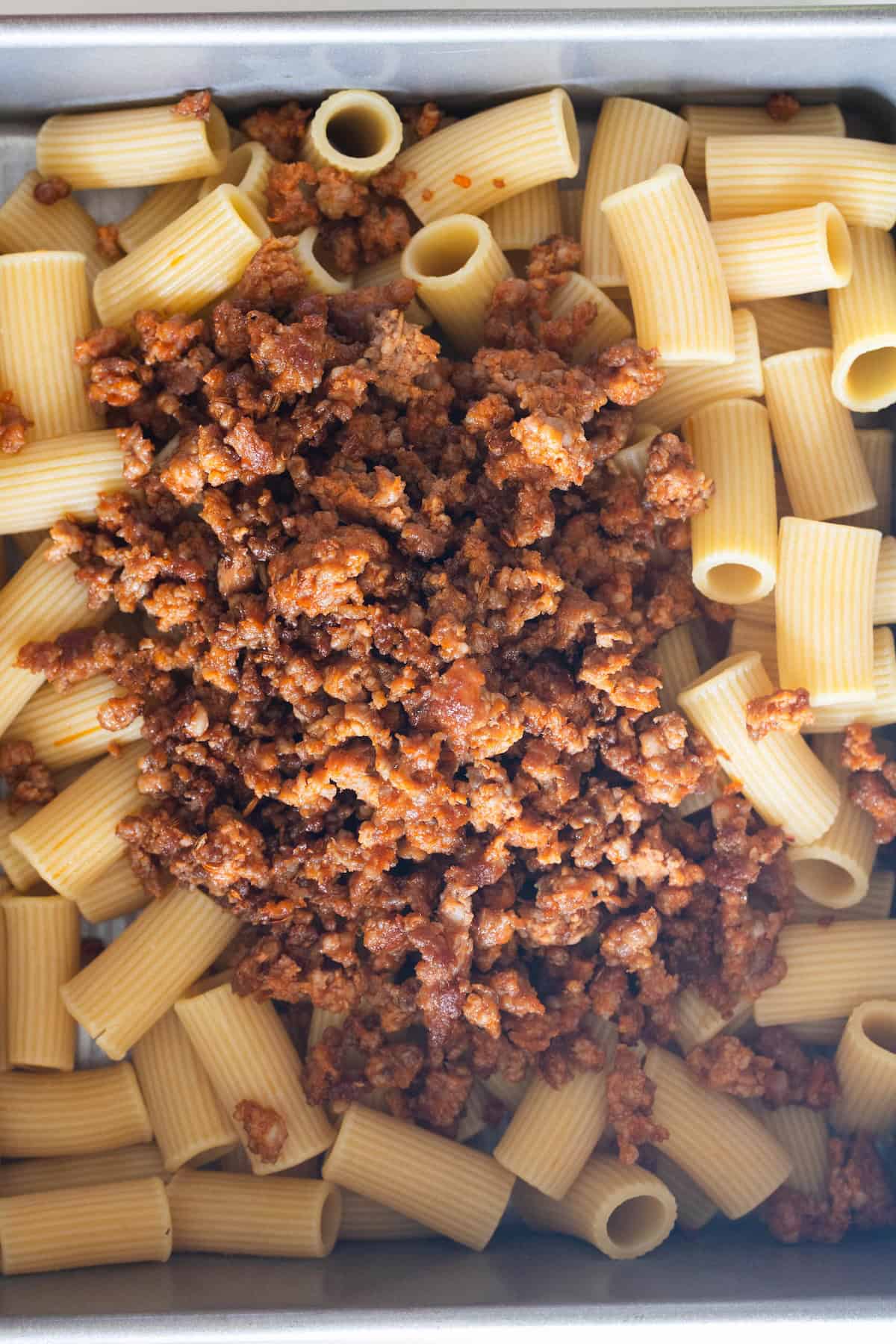 Cooked rigatoni pasta in a metal tray topped with crumbled ground meat in a red sauce, ready to be mixed or baked for a classic Cheesy Baked Rigatoni dish.