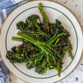 A plate of garlic broccolini arranged neatly on a round white dish with a green rim, placed on a speckled surface next to a blue and white towel.