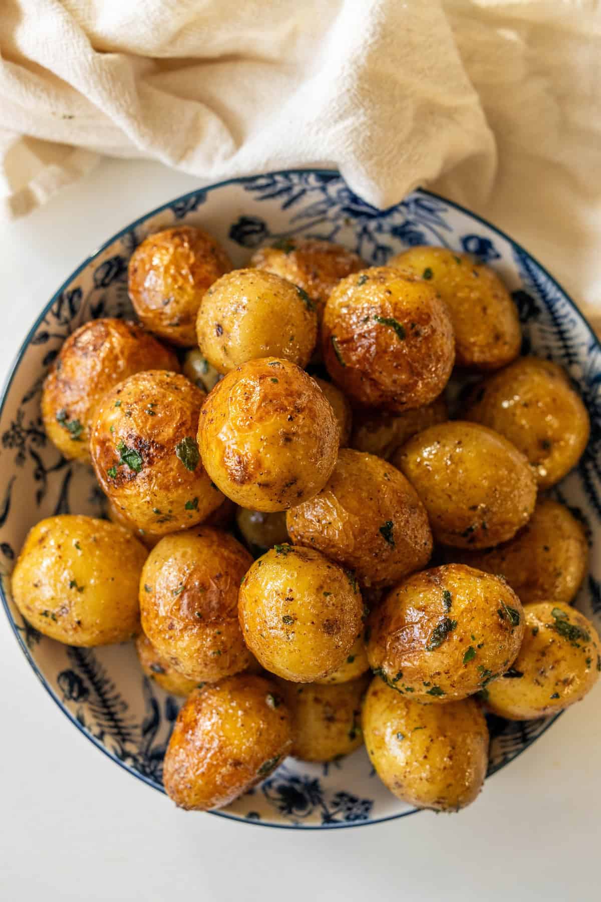 A bowl of butter poached potatoes garnished with herbs, placed on a white surface with a beige cloth in the background.